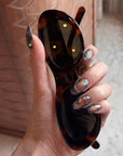 Hand holding oversized sunglasses while wearing black, gray, and copper handmade press-on nails with metallic fan patterns.