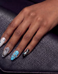 Hand wearing Artisian Orbit press-on nails with white, silver, blue, and black accents resting on a black leather surface