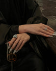 Tabletop lifestyle image of a hand model wearing Baroque Crown press-on nails with silver chrome, pearls, rhinestones, and white baroque accents