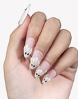 White background hand model wearing Boo Cuties press-on nails with pale pink bases, raised white ghost tips, and black eye details