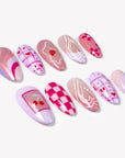 Angled flat lay of Candy Crush press-on nails with pink and purple tones, heart grid patterns, cherries, flowers, swirls, and white line art details