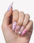 Hand model wearing Candy Crush press-on nails with pink and purple tones, heart grid nail art, strawberry detail, white line art, and flower accents