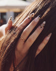 Hand model touching hair while wearing Chestnut Bloom press-on nails with nude pink bases, brown floral petals, black French tips, and gold accents