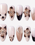 Chestnut Bloom press-on nails with nude pink bases, brown floral petals, black French accents, rhinestones, and glossy finish