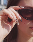Close-up hand model wearing Chestnut Bloom press-on nails with brown floral petals, black French tips, and rhinestone accents beside sunglasses