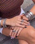 Seated hand model wearing Dark Masquerade press-on nails with glossy black tips, pink fishnet designs, rhinestone accents, and silver jewelry