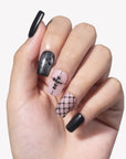 White background hand model wearing Dark Masquerade press-on nails with glossy black nails, black glitter accents, pink rhinestone cross, and fishnet pattern