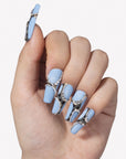 White background hand model wearing Denim Luxe press-on nails with sky blue bases, gold black leopard accents, pearl chains, and rhinestones