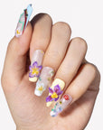 Hand model wearing Floral Symphony press-on nails with 3D purple flowers, white blossoms, mushrooms, and rhinestones
