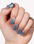 Hand model wearing Frosted Cherry press-on nails in silver and blue with red gem accents on a white background
