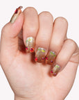 Hand model wearing Frosted Cherry press-on nails in gold and green glitter with red gem accents on a white background