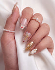 Hand model wearing blush pink and gold jeweled press-on nails with pearl rings on a white draped background.
