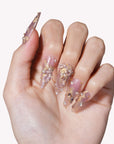 Close-up of a hand model wearing Gilded Petal Waltz press-on nails in soft lavender and gold with floral accents, pearls, and rhinestones