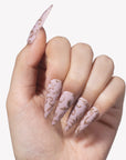 Close-up hand model wearing Gilded Petals stiletto press-on nails with soft pink bases and raised gold vine patterns