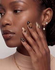 A hand model wearing warm brown and gold press-on nails poses beside the face in a close-up portrait.