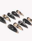 Angled flat lay of Gothic Elegance press-on nails with matte black and nude dot designs, silver cross charms, black rhinestones, lace effects, and butterflies