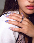 Fashion close-up of a model wearing Iris Bloom iridescent purple floral press-on nails with silver rings