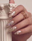 Close-up of a hand model wearing white and gold stained-glass floral press-on nails beside a stone column prop.