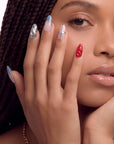 Face close-up hand model wearing Liquid Mercury press-on nails with silver chrome swirls, blue gray tips, and a red textured accent nail