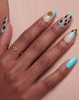 Close-up of a hand wearing turquoise and cream almond press-on nails with black abstract spots and a gold flower accent