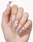 Hand model wearing Luxurious Treasure press-on nails with nude pink square bases and large pink and clear rhinestone gems on a white background