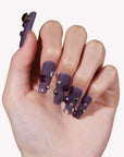 Hand model wearing Luxurious Treasure press-on nails in a muted purple square style with dark purple, black, and clear rhinestone gems