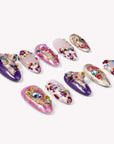 A flat lay of ten almond-shaped press-on nails on a white background featuring deep purple and pearly pink finishes, decorated with colorful rhinestones, pearls, 3D purple bows, and molten gold metallic accents