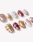 Angled flat lay of Molten Mirage press-on nails with amber and purple jelly swirls, champagne shimmer, gold shell accents, rhinestone trim, and raised gem details