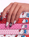 Hand model wearing Monochrome Meow press-on nails with black and white cat designs, silver bows, pearl charms, polka dots, and a colorful cartoon box background