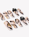 Scattered flat lay of beige press-on nails with black swirls, gold accents, and floral embellishments.