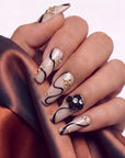 Close-up of a hand wearing beige press-on nails with black swirls, gold accents, and floral embellishments.