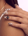 Side close-up of a hand wearing beige press-on nails with black swirls, gold accents, and floral embellishments.