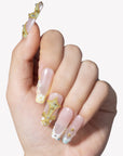 Hand model wearing Opal Dynasty press-on nails with nude pink bases, gold crown accents, pearl flower details, rhinestones, and blue translucent tips