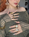 Hand model wearing Opal Dynasty press-on nails with nude pink bases, gold crown details, pearl accents, rhinestones, and layered gold jewelry