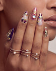 Close-up of a hand model wearing Opulent Ribbon Reverie press-on nails in purple pink and ivory with gold ribbon details and colorful gem accents