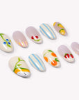 Set of handmade press-on nails with blue stripes, cherries, lemons, strawberries, and peach designs on a white flat lay.