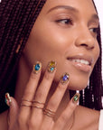 Close-up of a hand model wearing multicolor gemstone press-on nails with gold rings and a neutral background.