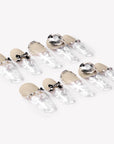 Angled flat lay of Rose Gold Whispers press-on nails with cream nude tops, translucent white floral tips, bow charms, rhinestone chains, and large crystals