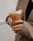 Lifestyle hand model in a beige blazer holding a drink while wearing Rosegold Flutter press-on nails with rosegold butterfly and rhinestone details