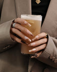 Hand model holding a drink while wearing Rosegold Flutter press-on nails with glossy nude, white, and burgundy designs plus rosegold butterfly accents