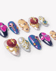 Angled flat lay of Saint of Desire press-on nails with pink flowers, red heart charms, gold cross details, pearls, and gemstones
