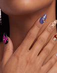 Hand model wearing Saint of Desire press-on nails with pink flower, red heart charm, gold cross, pearls, and gemstone accents