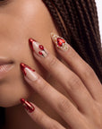 Close-up of a hand model wearing Scarlet Heart Oath press-on nails with red metallic tips, nude bases, hearts, and gold bead accents