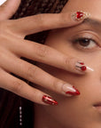 Lifestyle portrait of a hand model wearing Scarlet Heart Oath press-on nails with red metallic tips, nude bases, and gold bead heart accents