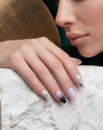 Face close-up hand model wearing Stellar Muse press-on nails with black and silver star charms, rhinestones, silver beads, nude pink bases, and black gradient accents