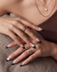 Hand model wearing Timeless Serenity press-on nails with nude bases, black French tips, white 3D flowers, and gold jewelry against a brown top