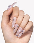 Hand model wearing translucent nude pink stiletto press-on nails with pearl clusters, gold studs, and crystal accents