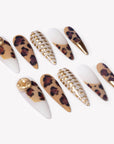 Angled flat lay of Wild Majesty press-on nails with tan leopard print, white bases, raised gold dot accents, gold charms, and metallic gold stiletto tips