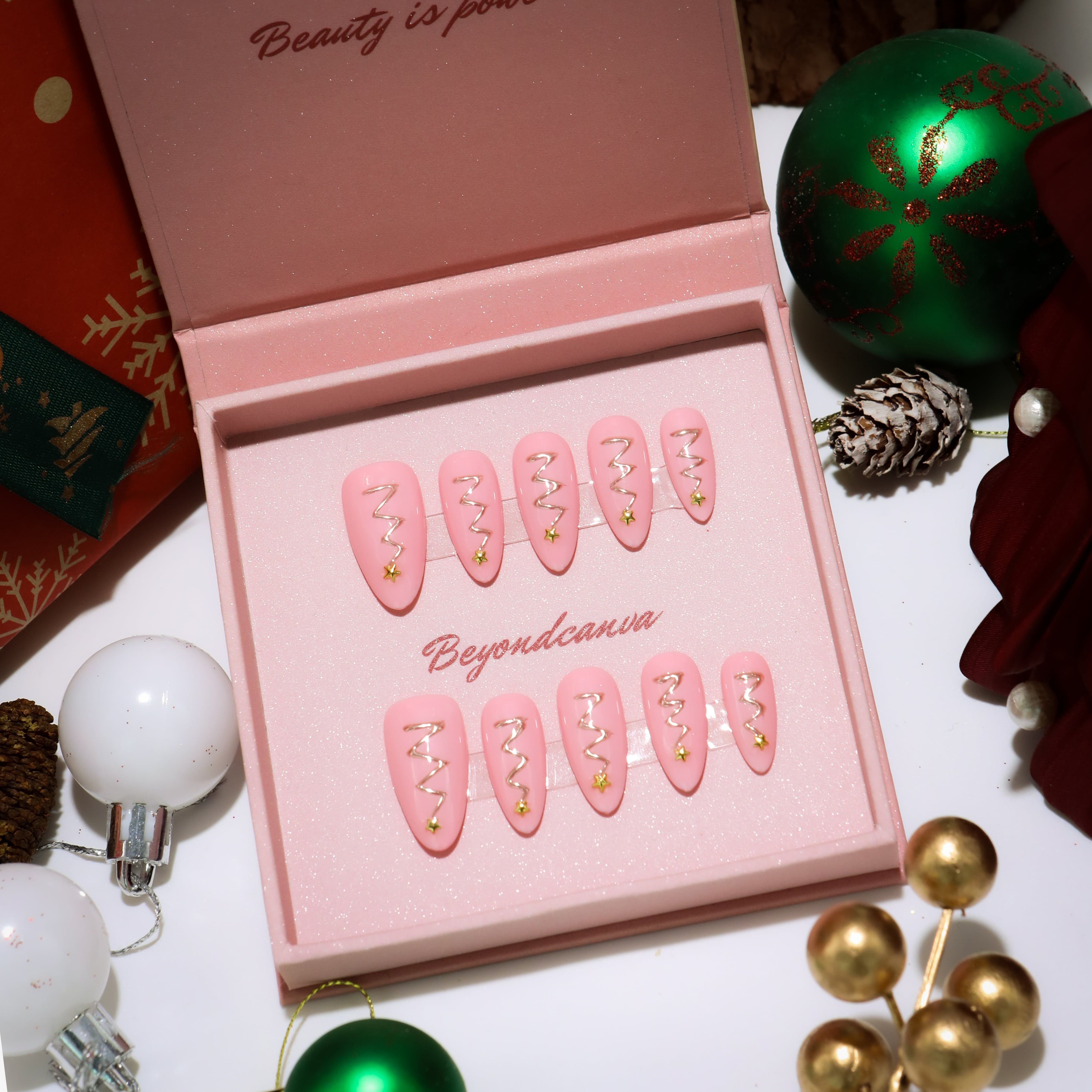 This beyondcanva press-on nail set features pastel pink nails with festive gold zigzag accents and star embellishments, resembling Christmas trees. Packaged in a luxurious pink box, it’s perfect for a chic holiday look.