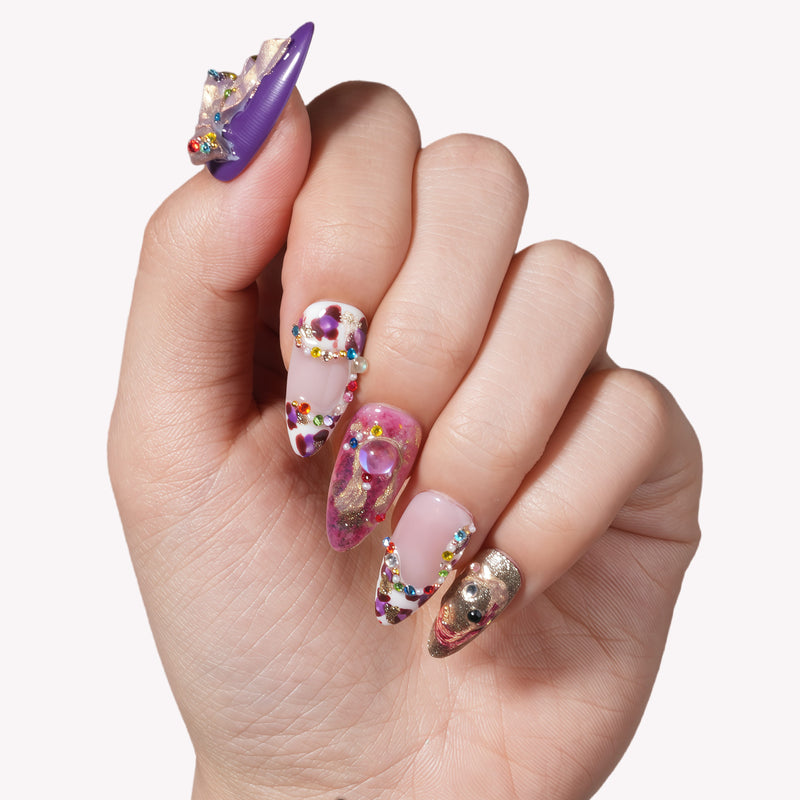 Close-up of a hand showcasing elaborate 3D nail art on long almond-shaped nails featuring purple and gold themes, jeweled French tips with colorful rhinestones, and 3D charms against a plain white background